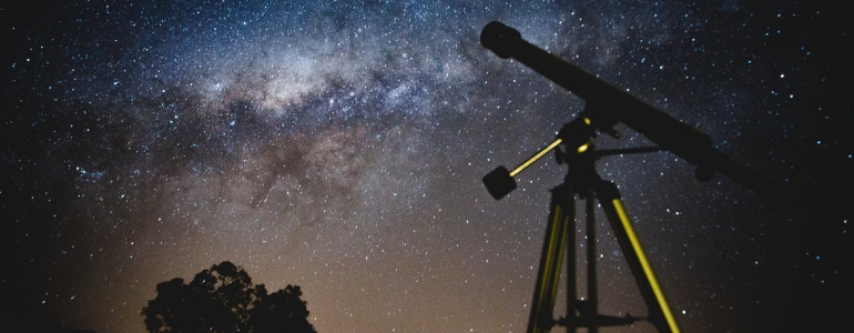 A telescope and a tree under the starry night sky with a flare.