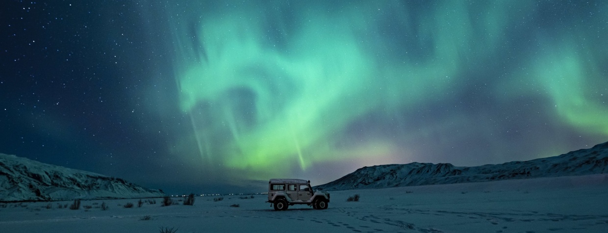 An SUV in a snowy field, against mountains and Northern Lights.