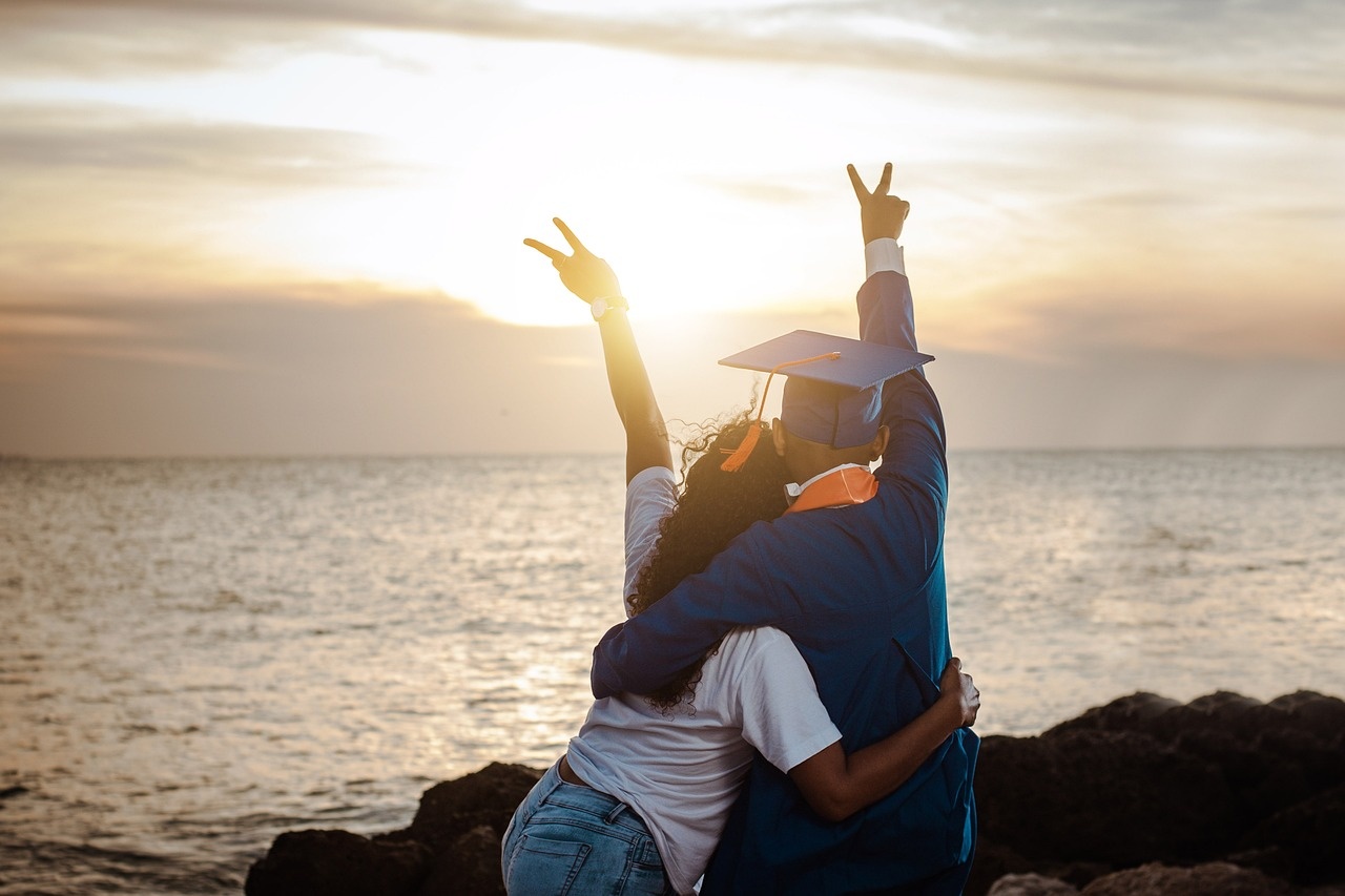 A graduate with close friend with raised arms on the seashore at sunset