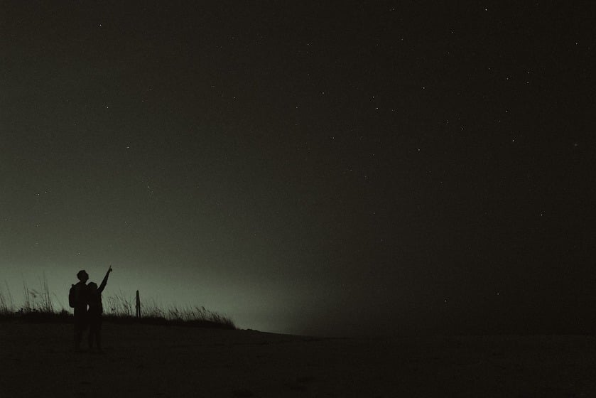 Two people standing in a field pointing at the dark sky where a few stars are shining