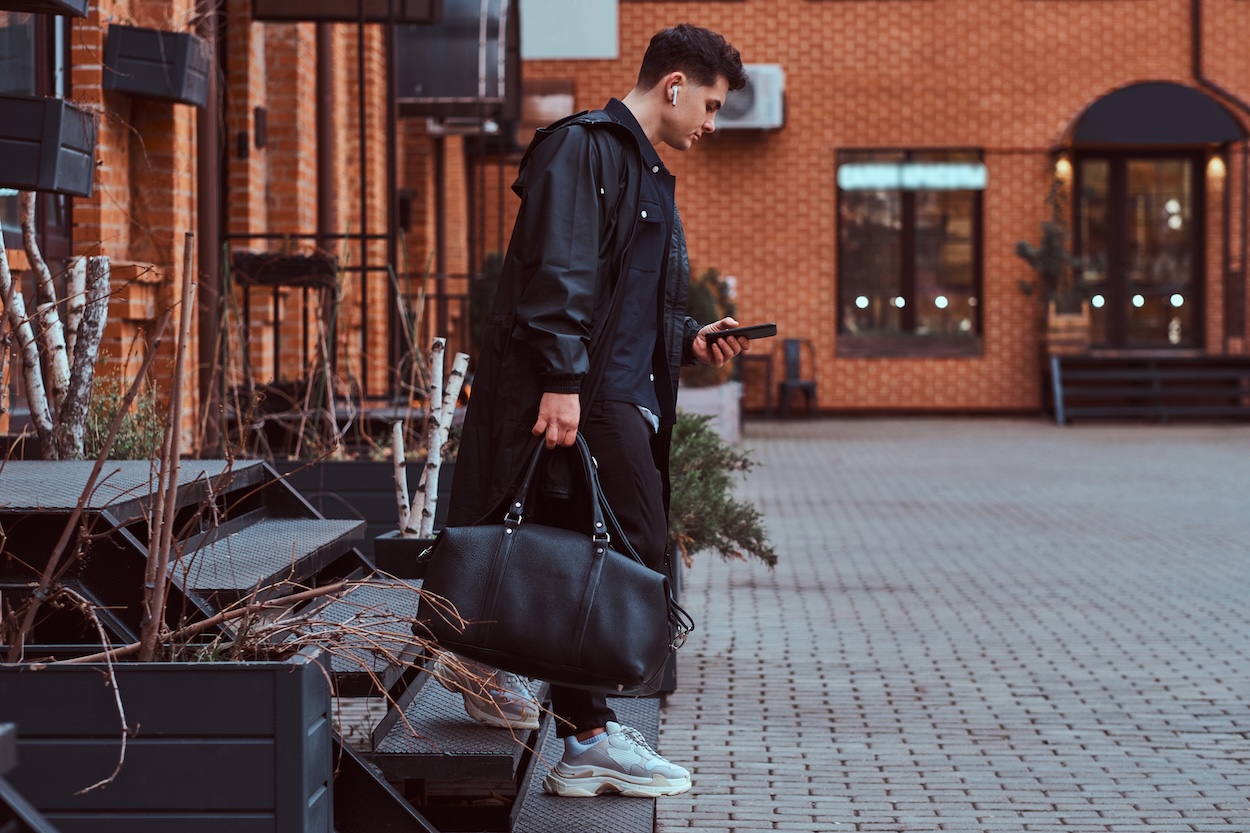 Un hombre joven vestido con un abrigo negro, llevando un bolso de viaje de cuero negro.