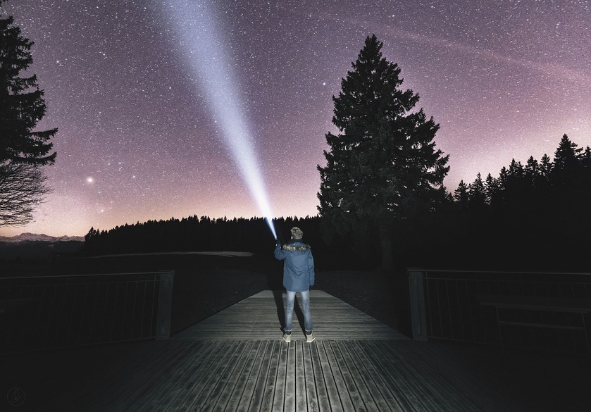 A person standing in the dark and pointing a flashlight at the night sky full of stars