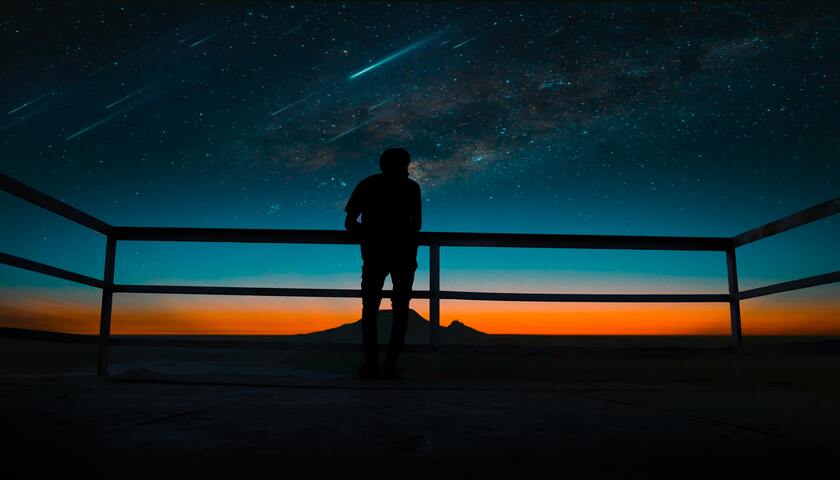 A person standing near a railing looking at shooting stars in a blue and orange sky