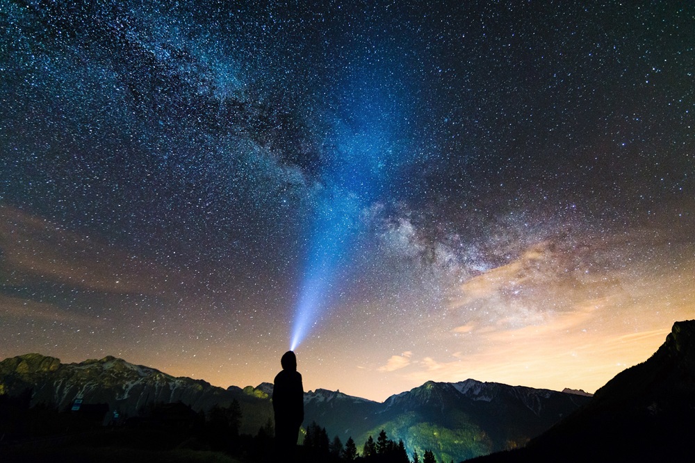 Silhueta de uma pessoa em pé perto das montanhas, olhando as estrelas no céu.