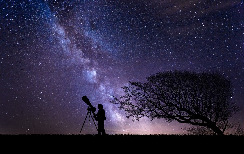 A person standing in front of colorful clouds and a crescent moon putting up a telescope